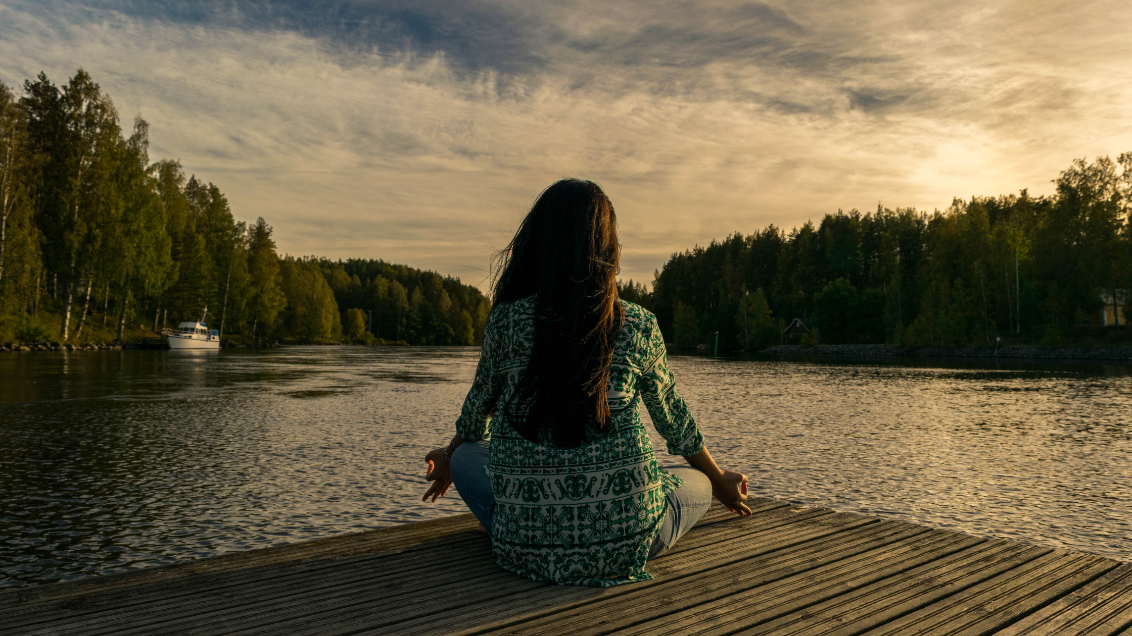 A person sits cross-legged on a wooden dock meditating by the lake at sunset, surrounded by trees and a boat in the background.